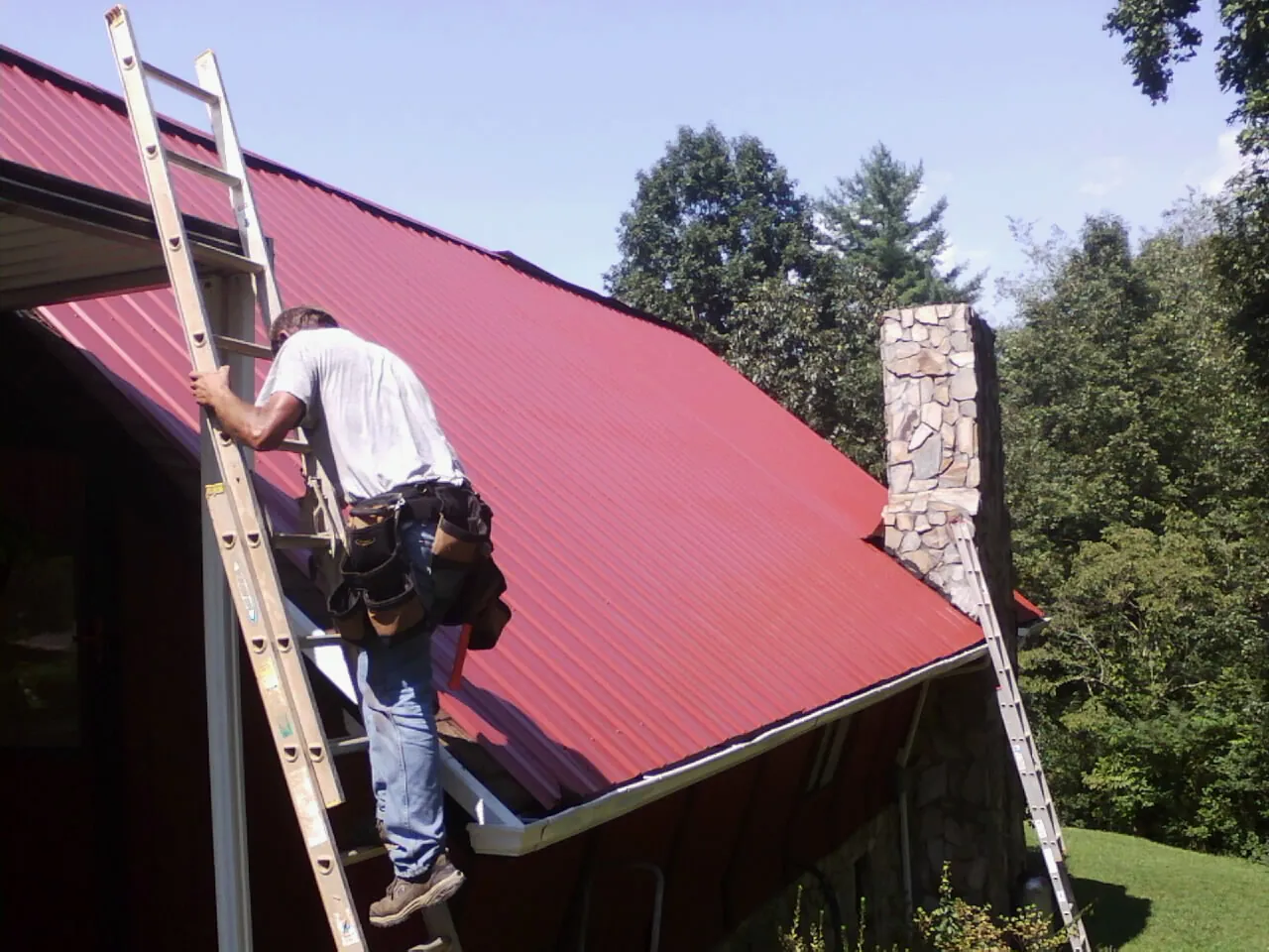 Roofer on a ladder beside a red metal roof and stone chimney — Soffit & Fascia Repair by Roofing Brothers in Haynesville
